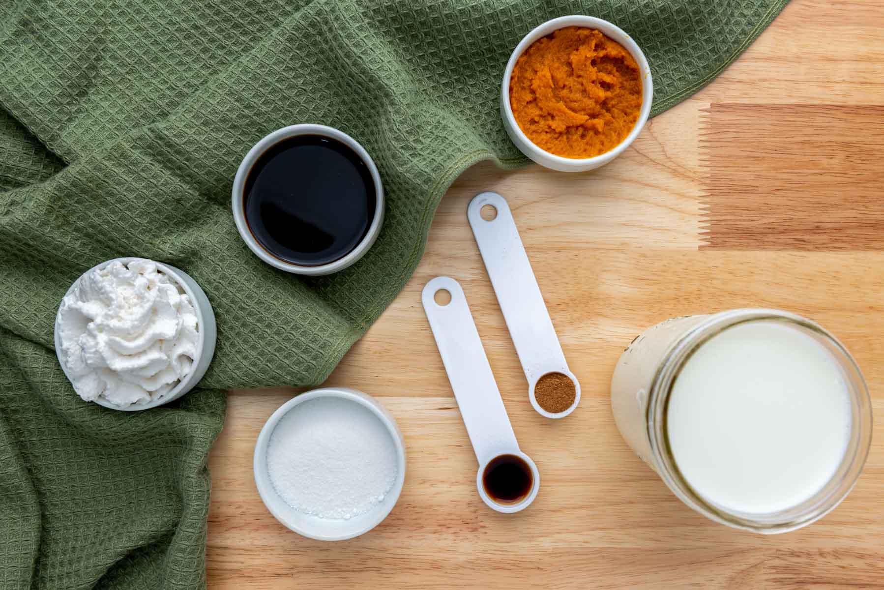 Ingredients on a wooden surface with a green cloth: pumpkin puree, whipped cream, milk, vanilla, spices, sweetener, and a dark liquid in bowls and measuring spoons.