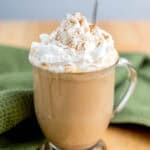 A clear glass mug of latte topped with whipped cream and a sprinkle of cinnamon sits on a wooden table next to a green cloth.