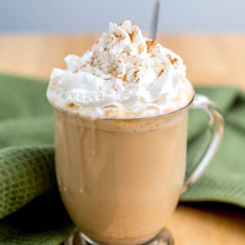 A clear glass mug of latte topped with whipped cream and a sprinkle of cinnamon sits on a wooden table next to a green cloth.