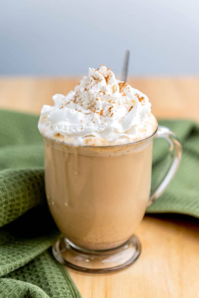 A clear glass mug of latte topped with whipped cream and a sprinkle of cinnamon sits on a wooden table next to a green cloth.
