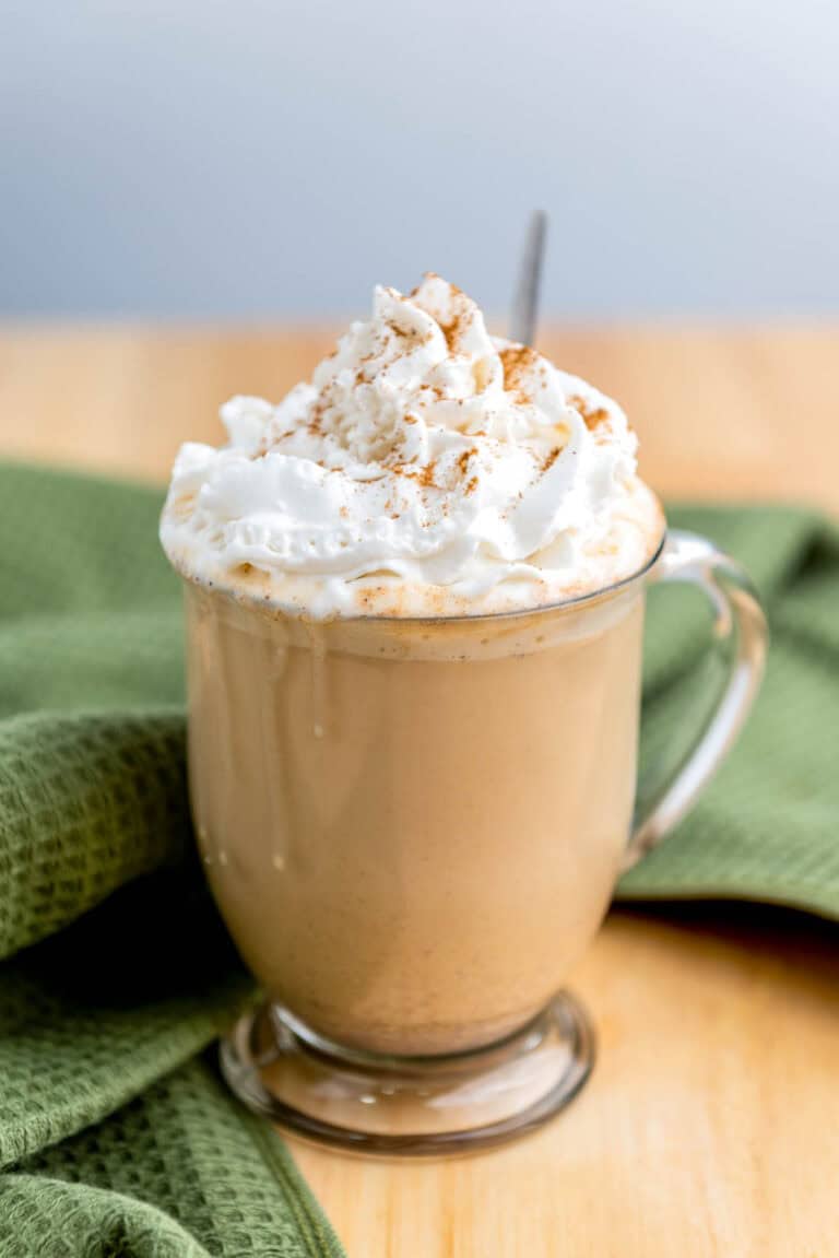 A clear glass mug of latte topped with whipped cream and a sprinkle of cinnamon sits on a wooden table next to a green cloth.