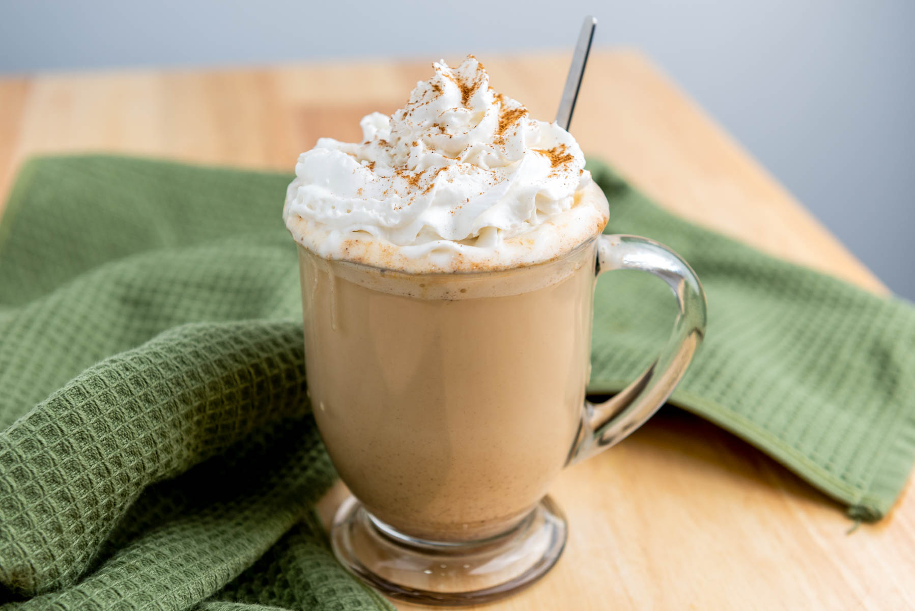 A glass mug of latte topped with whipped cream and sprinkled cinnamon sits on a wooden table with a green cloth.