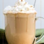 Glass mug filled with pumpkin spice latte topped with whipped cream and cinnamon, placed on a green cloth with a light background.
