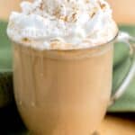 A glass mug of pumpkin spice latte topped with whipped cream and sprinkled cinnamon sits on a table with a green cloth behind it.
