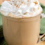 A glass mug of pumpkin spice latte topped with whipped cream and sprinkled with cinnamon, placed on a green cloth.