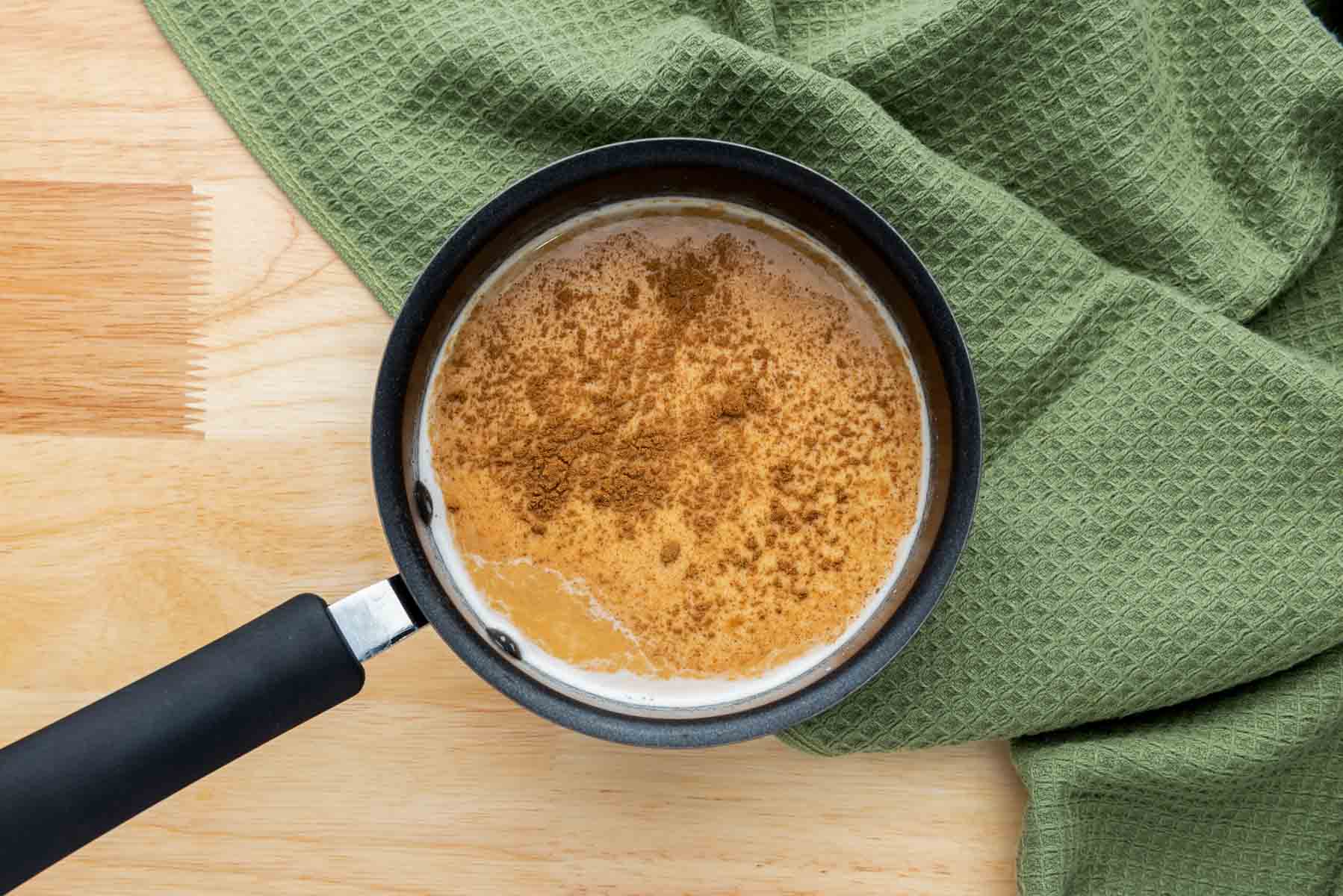 A saucepan with a creamy brown liquid and a layer of cinnamon on top sits on a wooden surface next to a green textured cloth.