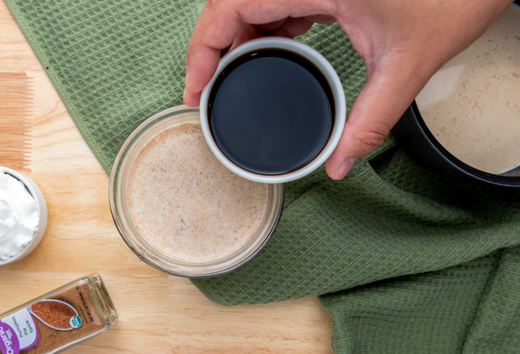 A hand holds a small bowl of dark liquid above a jar of light brown mixture on a wooden surface with a green cloth and ingredients nearby.