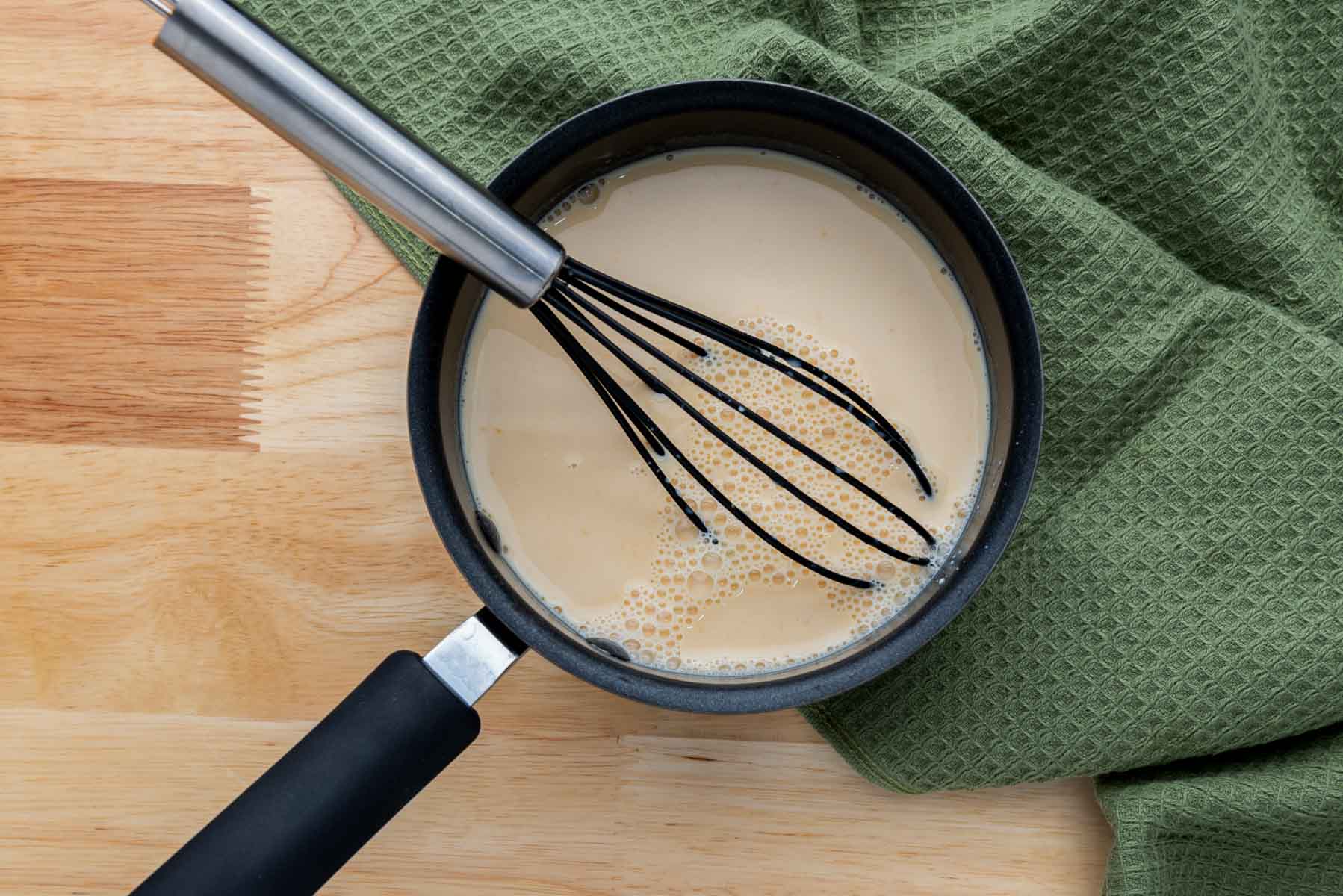 A black saucepan with a whisk inside contains a tan liquid, placed on a wooden surface next to a green textured cloth.