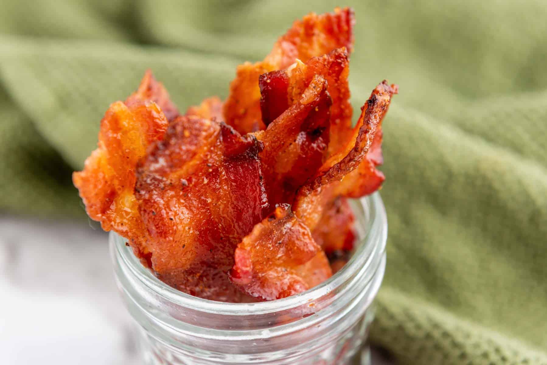 Crispy cooked bacon strips placed upright in a clear glass jar with a green textured cloth in the background.