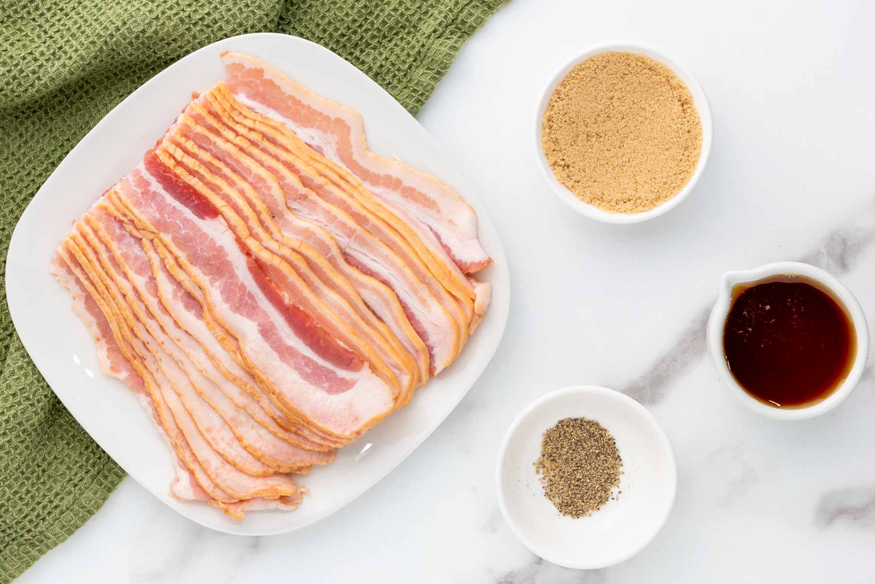 A plate of raw bacon strips next to bowls of brown sugar, ground black pepper, and maple syrup on a white marble surface with a green towel.