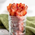 Crispy bacon strips arranged upright in a clear glass jar, placed on a marble surface with a green cloth in the background.