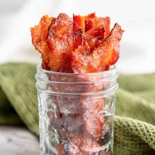 Crispy bacon strips arranged upright in a clear glass jar, placed on a marble surface with a green cloth in the background.