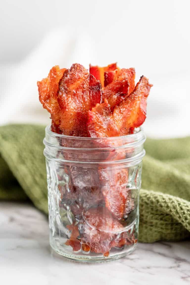 Crispy bacon strips arranged upright in a clear glass jar, placed on a marble surface with a green cloth in the background.
