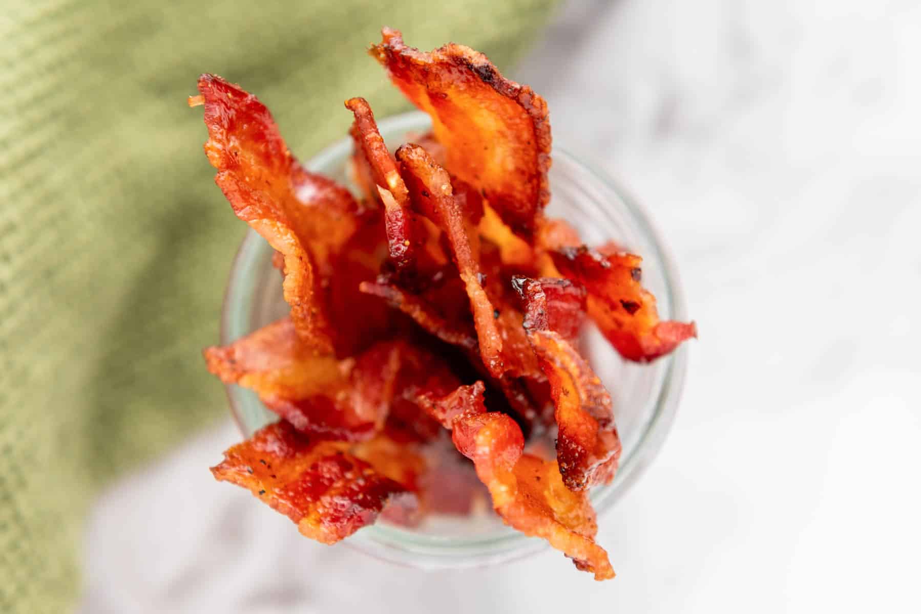 Crispy cooked bacon strips arranged upright in a clear glass on a white surface next to a green textured cloth.