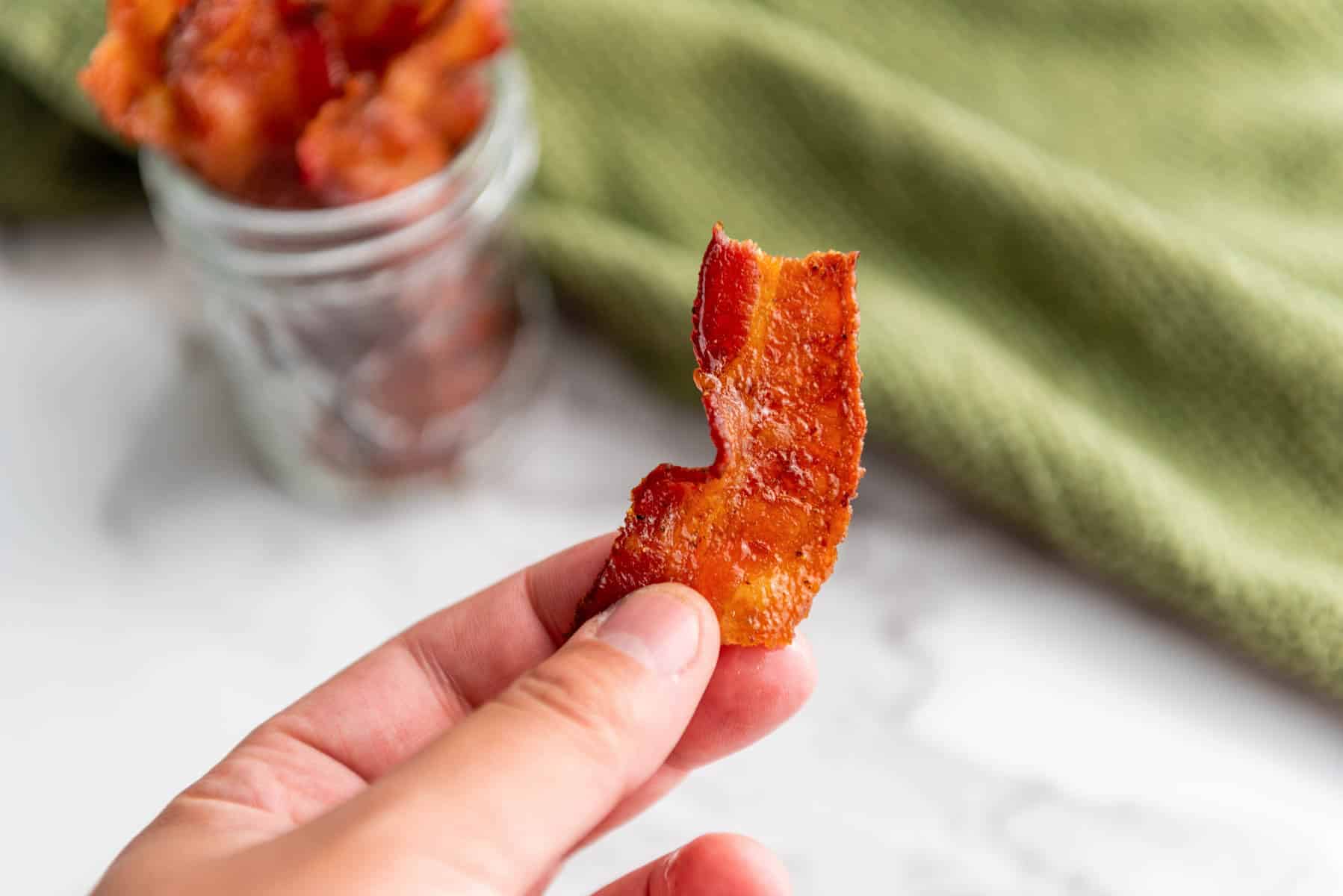 A hand holds a piece of crispy bacon with a jar of bacon pieces and a green textured cloth in the background.
