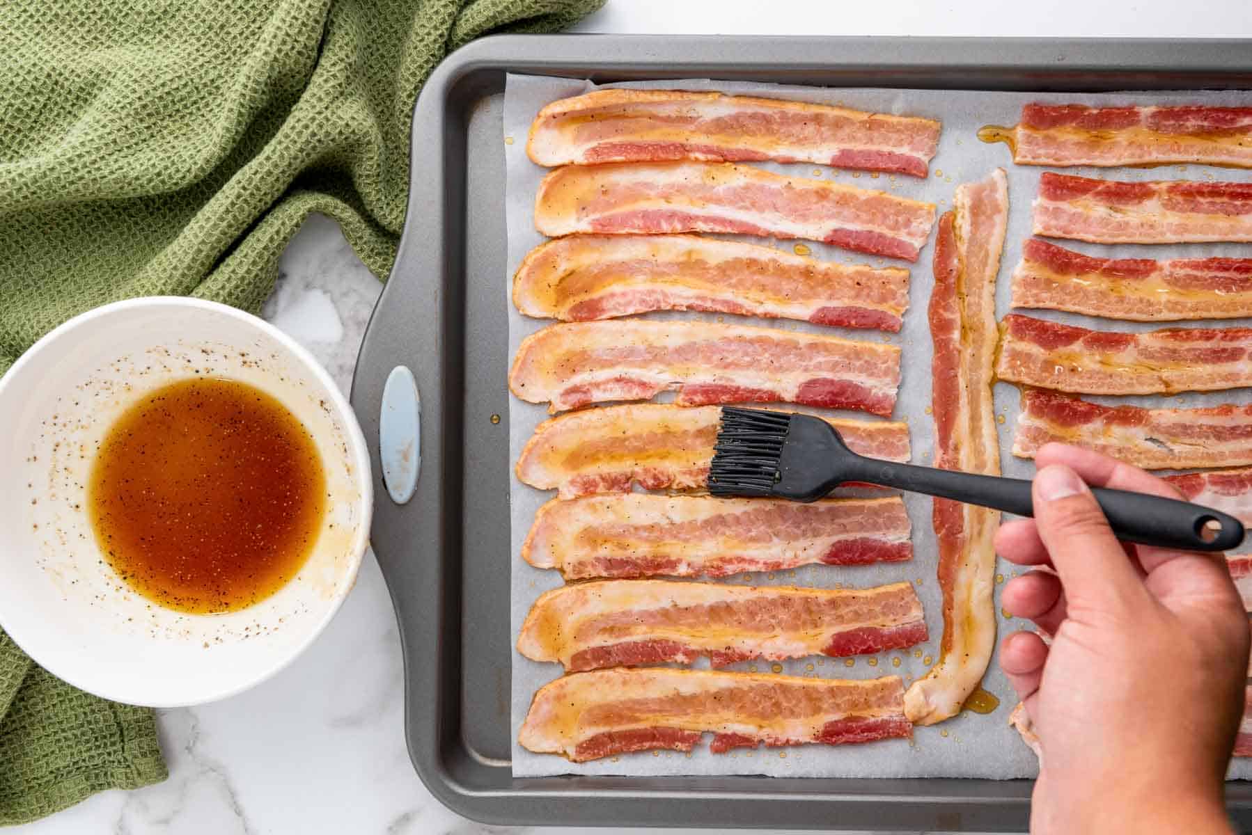 A hand uses a brush to apply a brown glaze to raw bacon strips arranged on a parchment-lined baking sheet, with a bowl of glaze nearby.