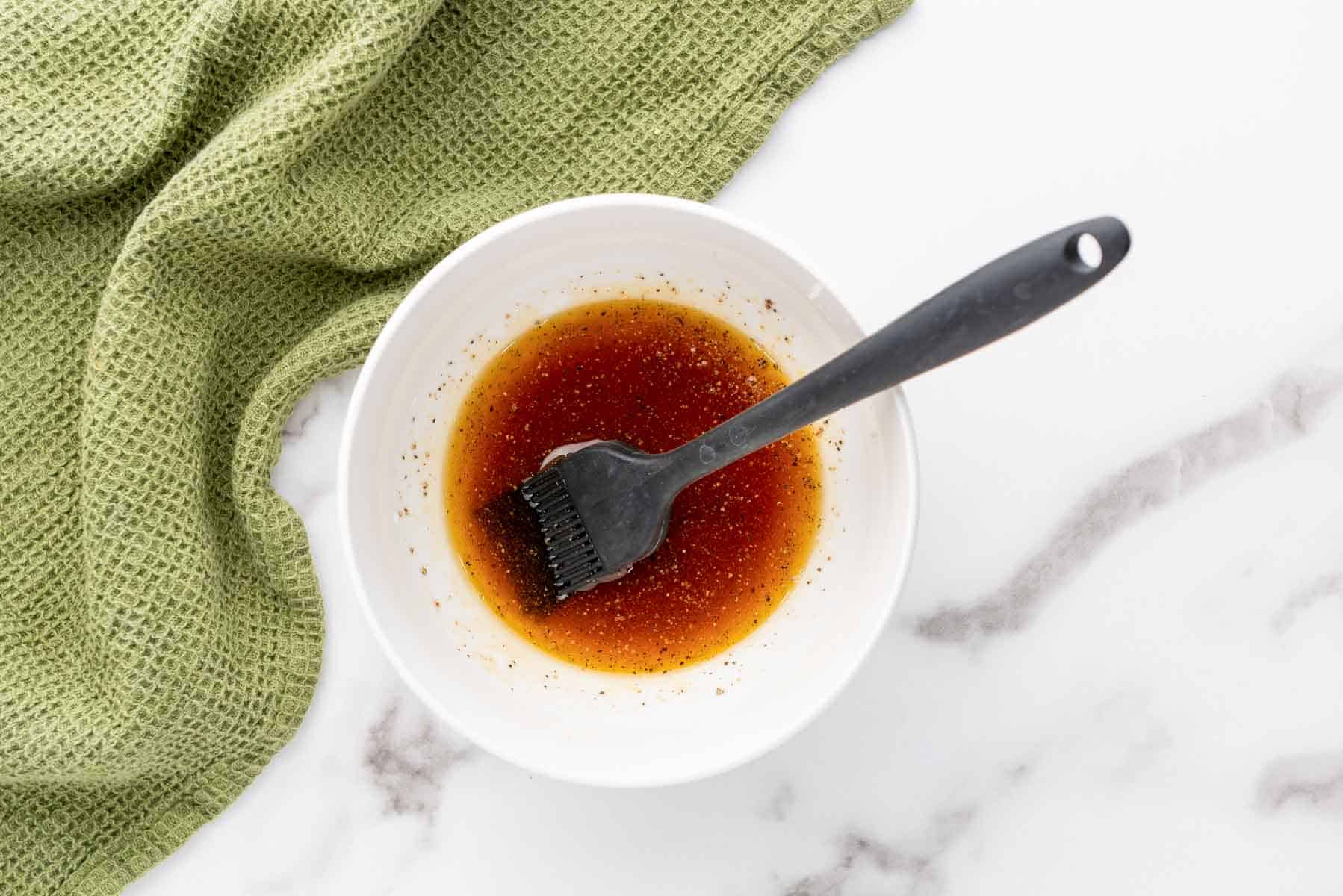 A white bowl with a brown marinade and a black basting brush on a marble surface, next to a green textured cloth.