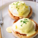 Two English muffin halves topped with perfectly poached eggs, rich hollandaise sauce, and a sprinkle of chives on a plate, with a fork and knife beside them.