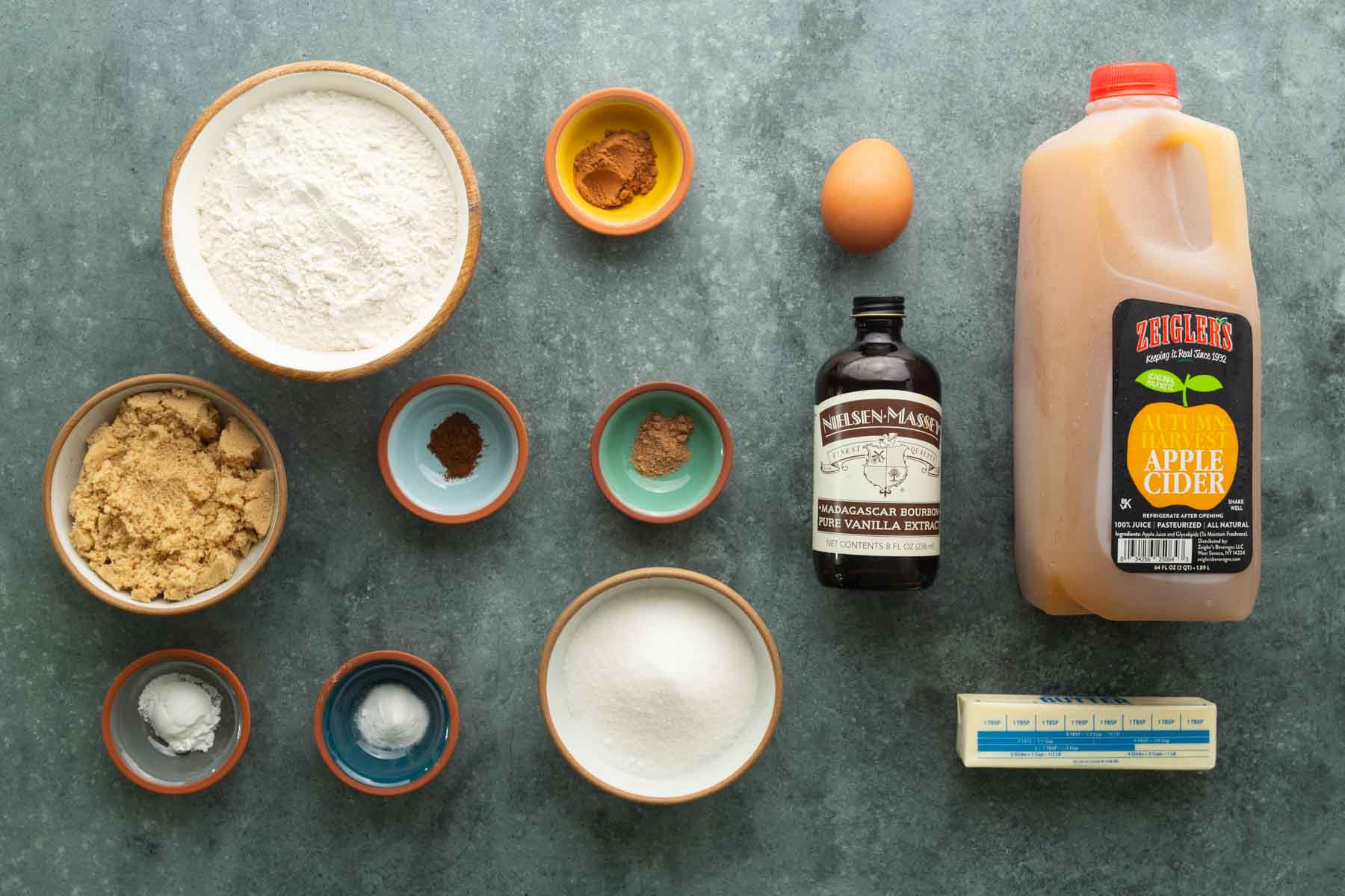 Ingredients for baking including flour, brown sugar, white sugar, a raw egg, apple cider, a stick of butter, vanilla extract, and spices in small bowls.