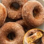 A bowl of cinnamon-sugar coated apple cider donuts, with an inset image showing the inside of a broken donut. Text overlay reads “Apple Cider Donuts”.
