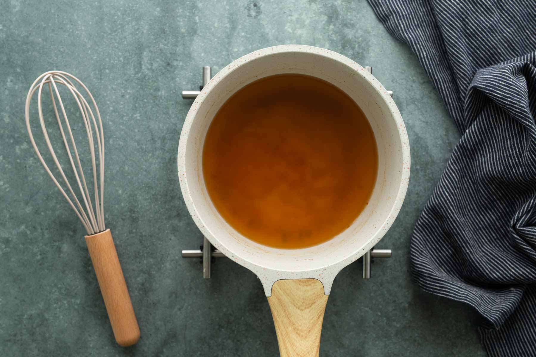 A light-colored saucepan containing amber liquid on a stove, with a wire whisk nearby.