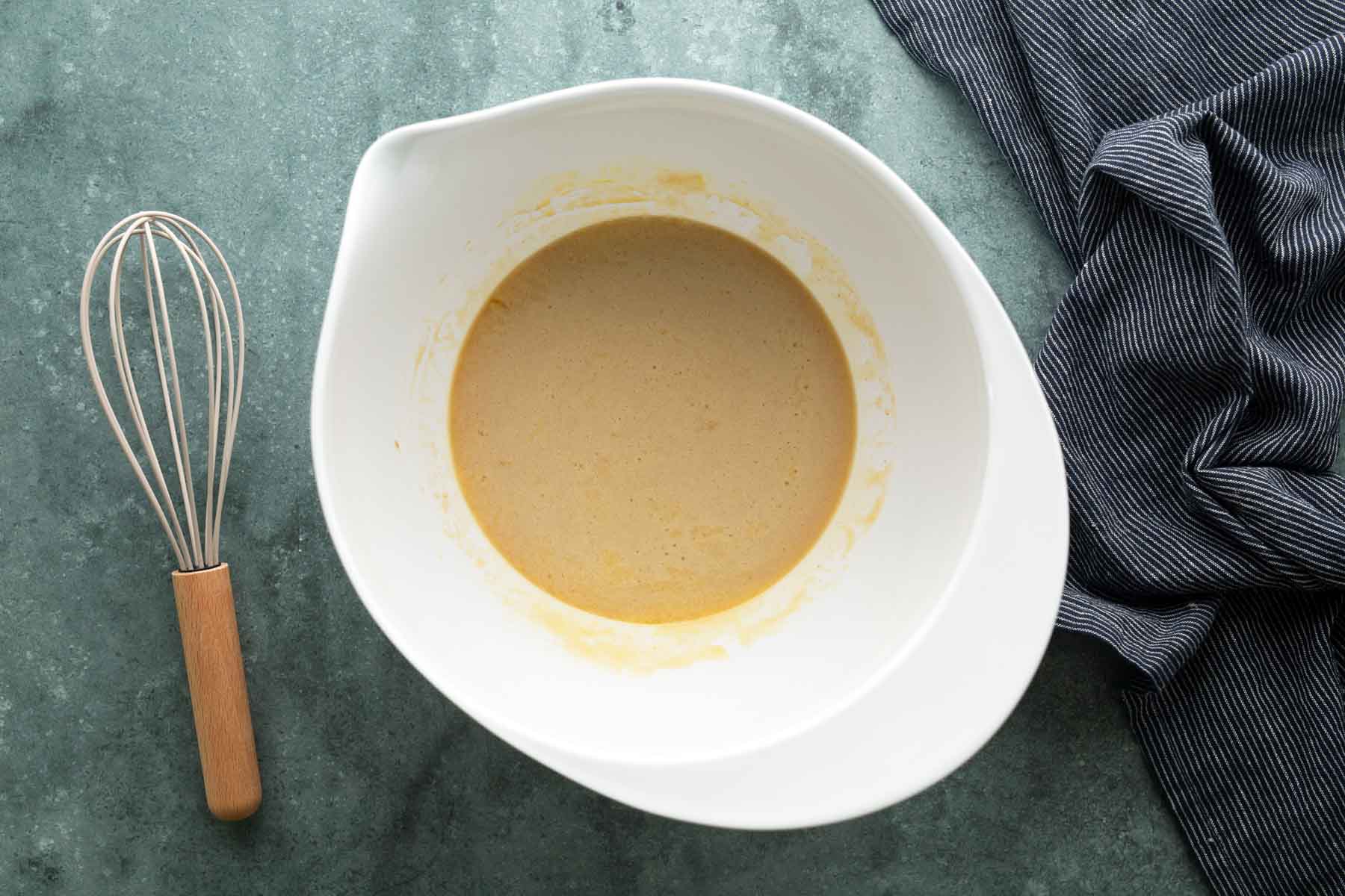 A white mixing bowl containing tan batter, with a wire whisk nearby.