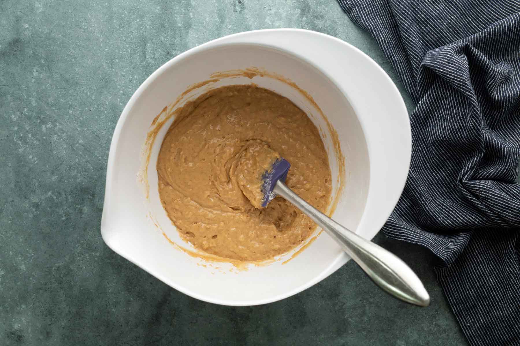 A white mixing bowl containing tan batter and a silicone spatula.