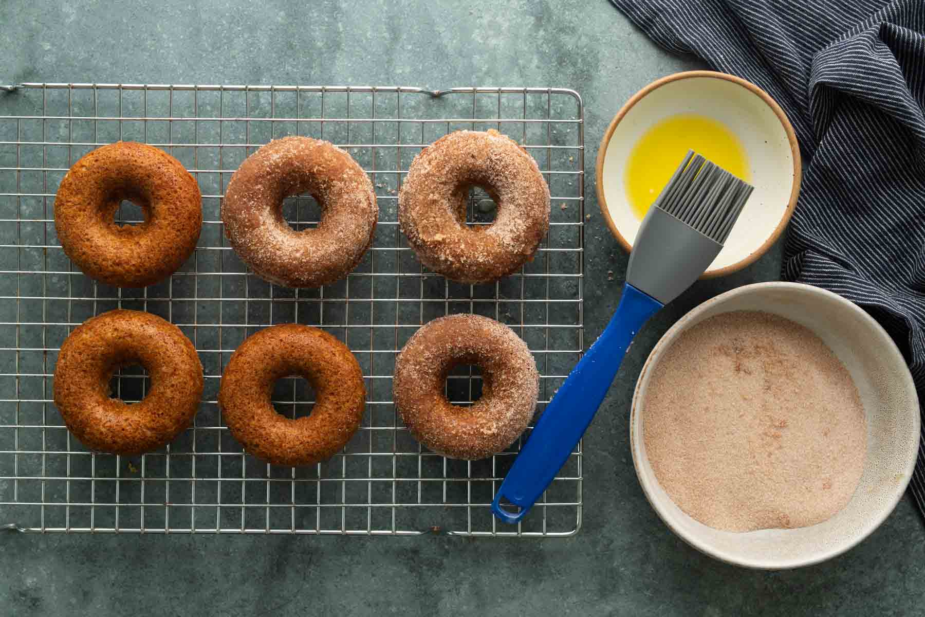 Six doughnuts on a wire rack next to a small bowl of melted butter with a silicone brush and a bowl of cinnamon sugar.