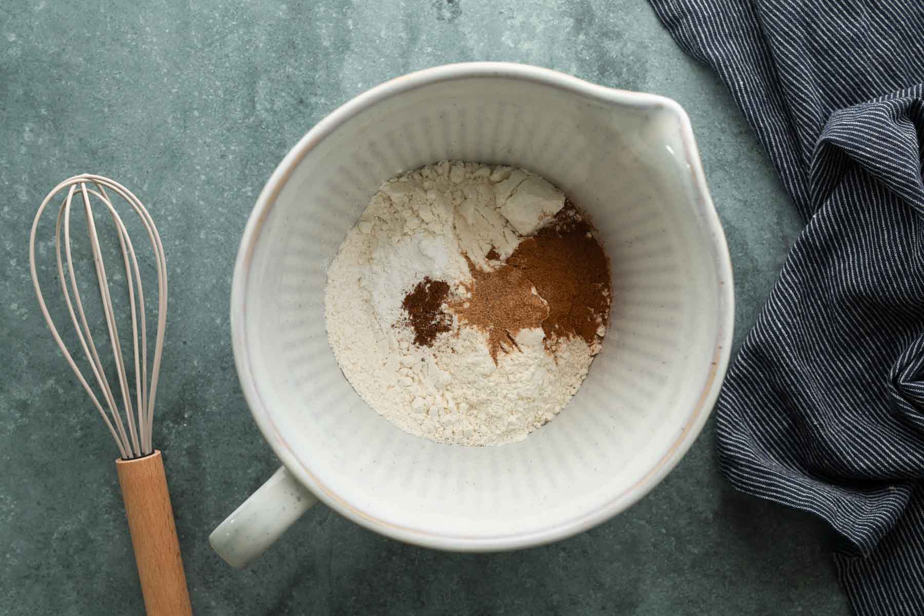 A mixing bowl containing flour and spices, with a whisk resting beside it on a blue surface.
